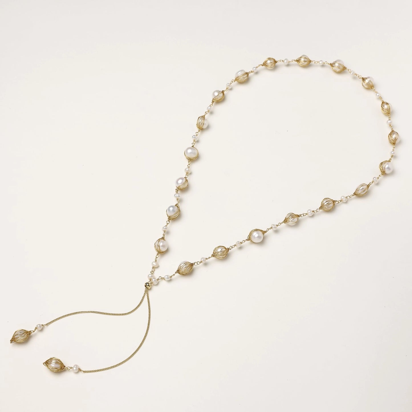Long gold-plated lariat necklace with wire-wrapped freshwater pearls and chain tassel detail on cream background.