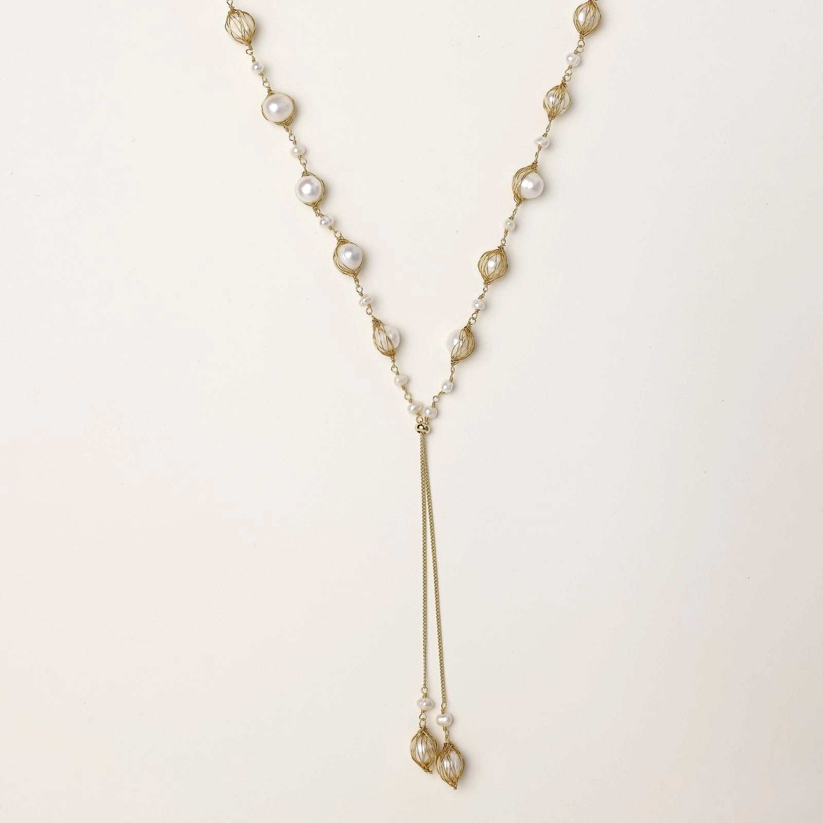 Long gold-plated lariat necklace with wire-wrapped freshwater pearls and chain tassel detail on cream background.