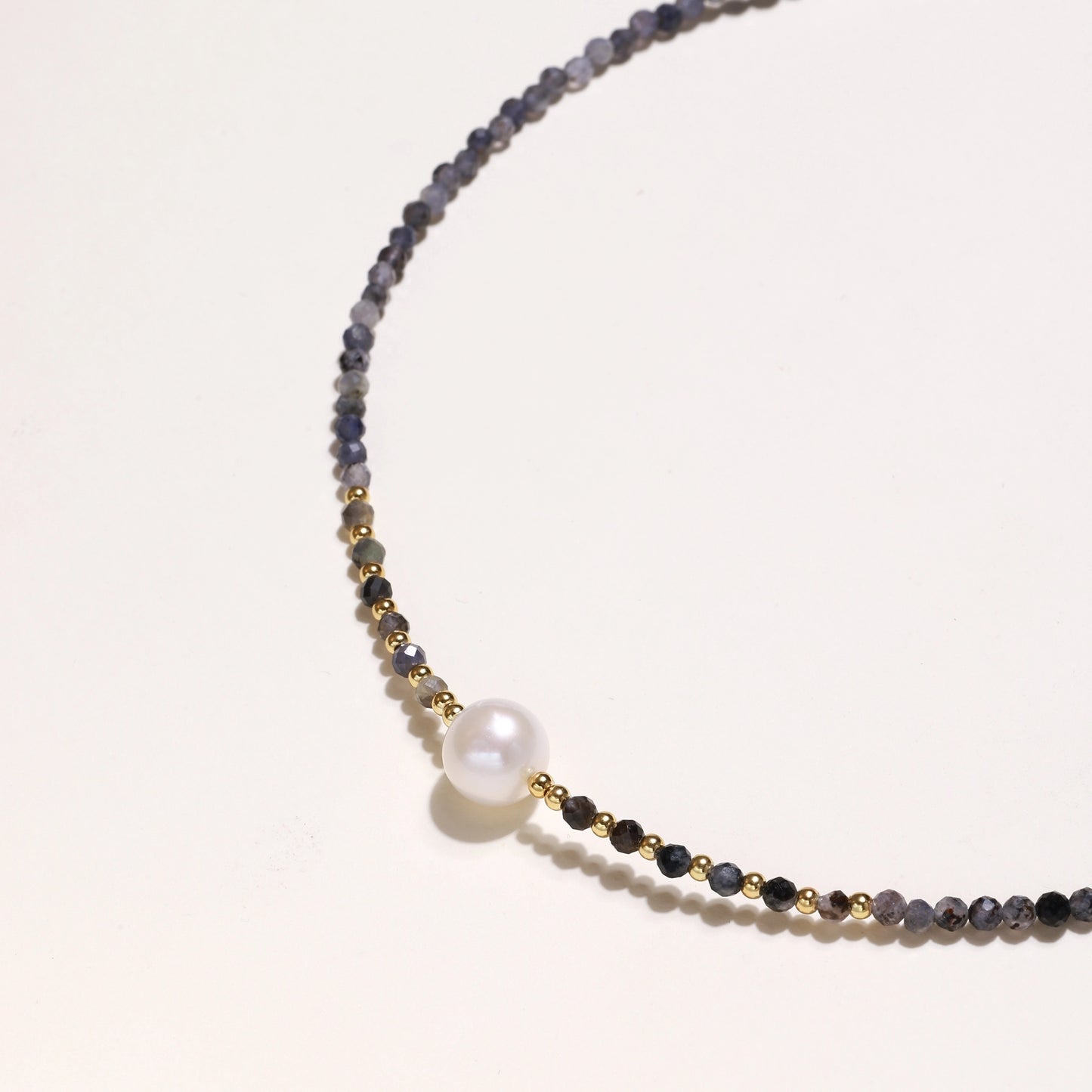 Delicate beaded necklace with violet iolite stones, gold spacers, and central white freshwater pearl on neutral background.