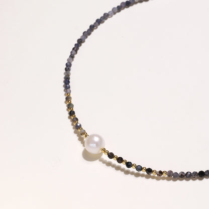 Delicate beaded necklace with violet iolite stones, gold spacers, and central white freshwater pearl on neutral background.