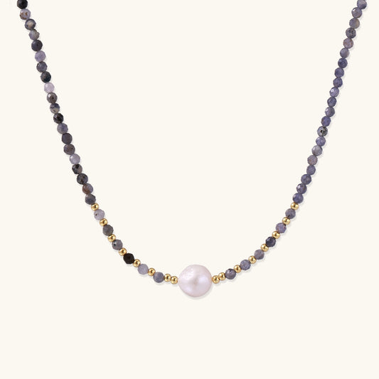 Delicate beaded necklace with violet iolite stones, gold spacers, and central white freshwater pearl on neutral background.