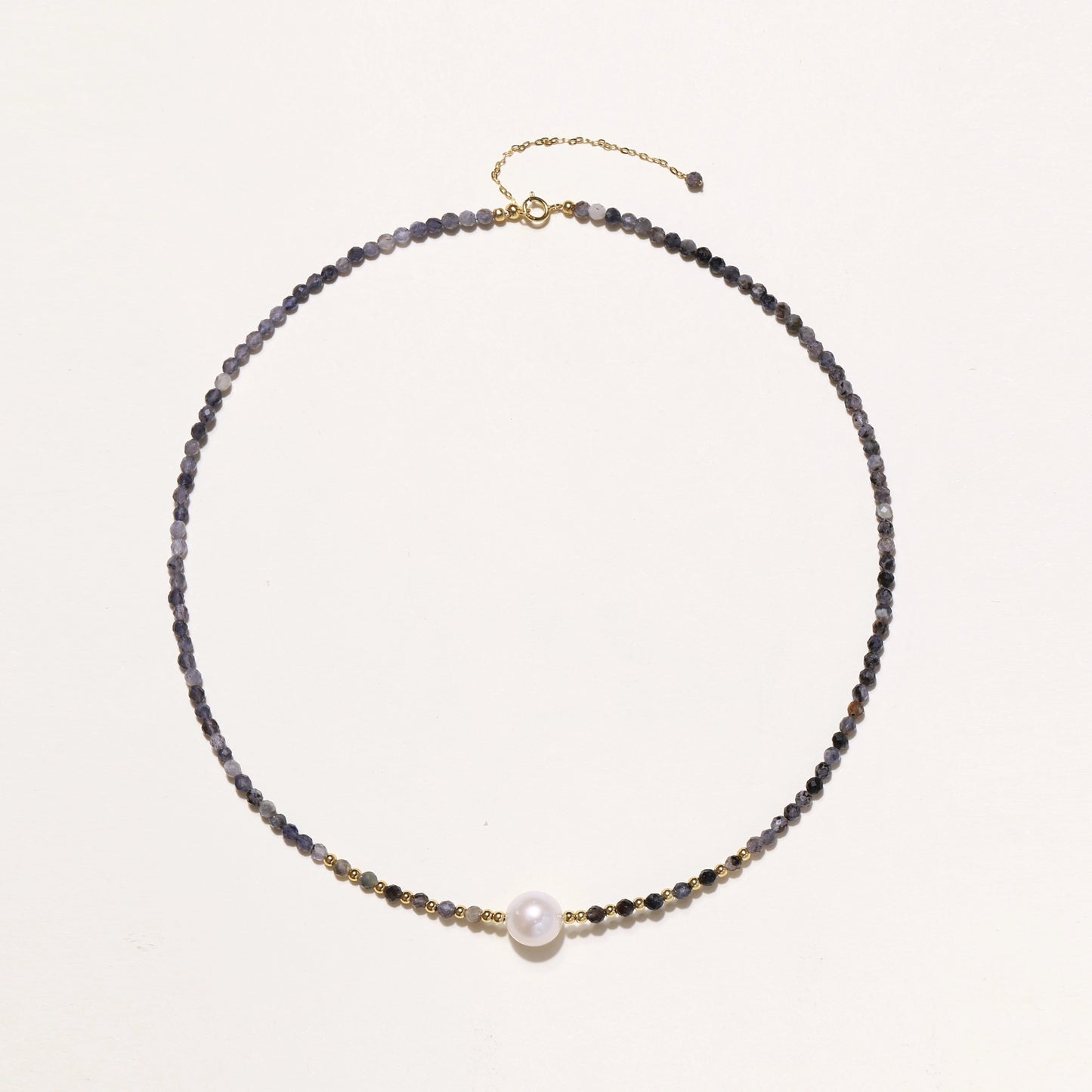 Delicate beaded necklace with violet iolite stones, gold spacers, and central white freshwater pearl on neutral background.
