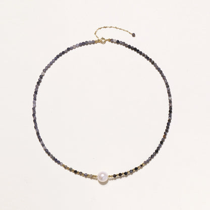 Delicate beaded necklace with violet iolite stones, gold spacers, and central white freshwater pearl on neutral background.