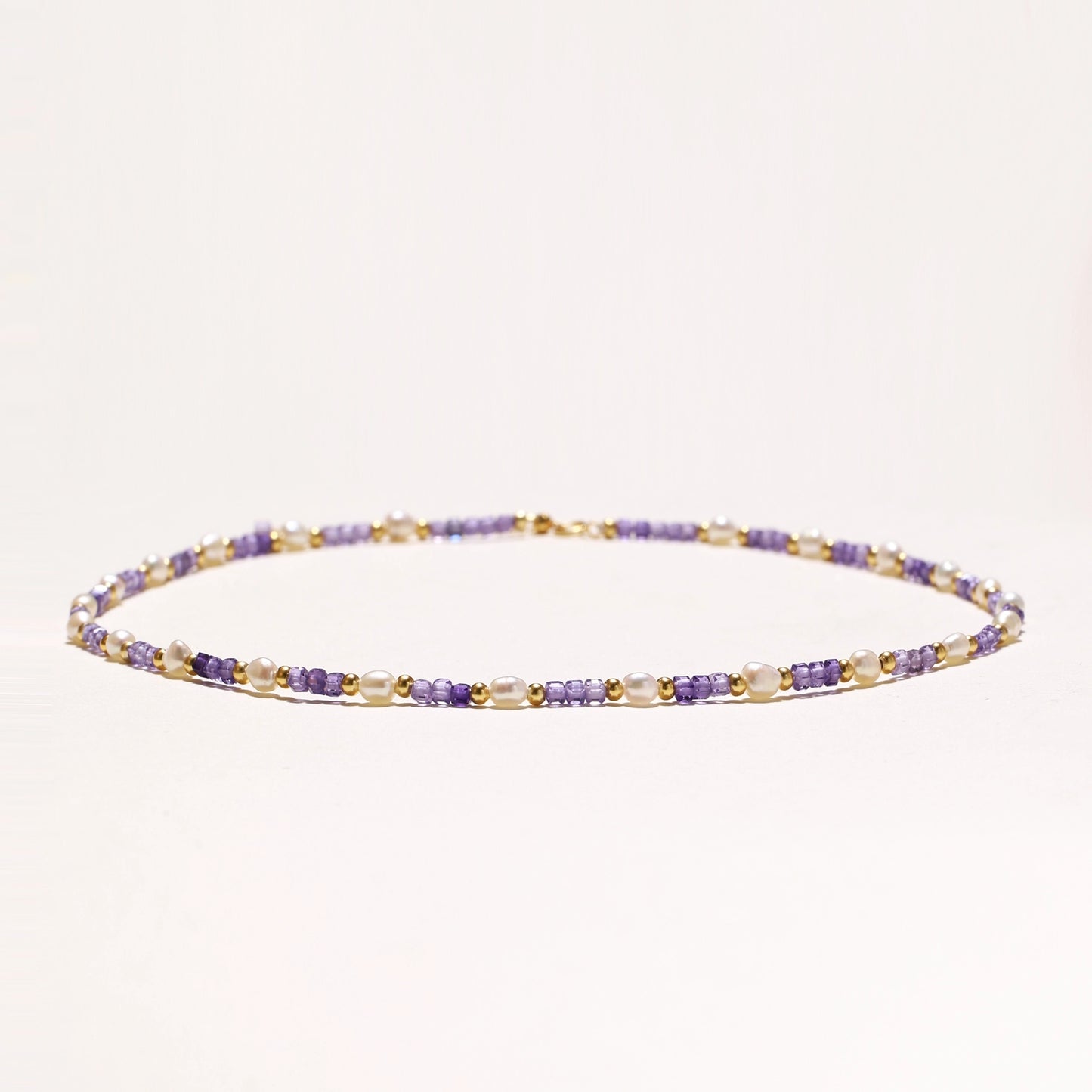 Delicate beaded necklace with alternating purple zirconia stones, white freshwater pearls, and gold-plated spacers on cream background.