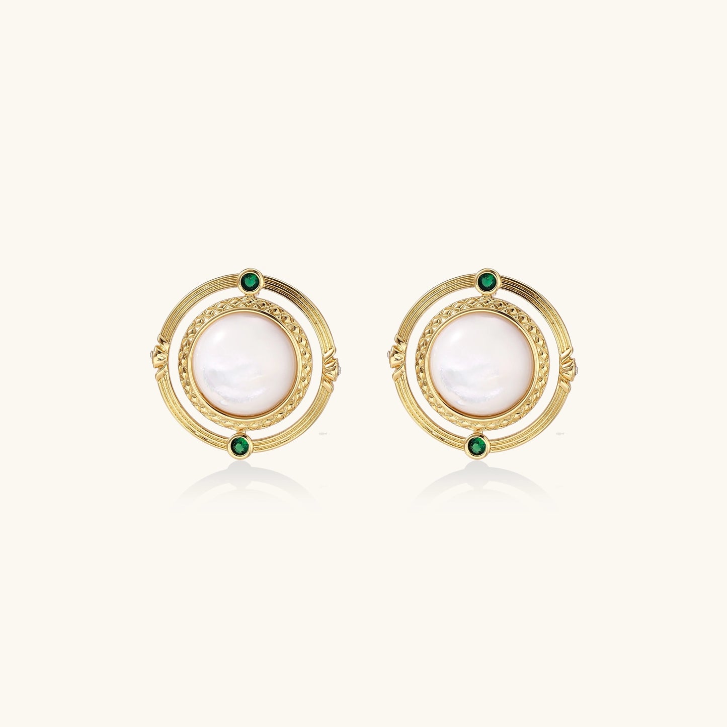 Close-up of gold-plated circular earrings with white mother-of-pearl centers and green zirconia accents on cream background.