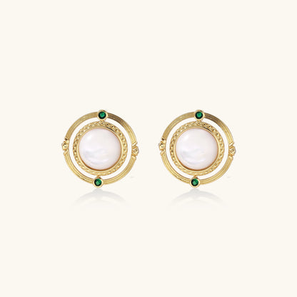 Close-up of gold-plated circular earrings with white mother-of-pearl centers and green zirconia accents on cream background.