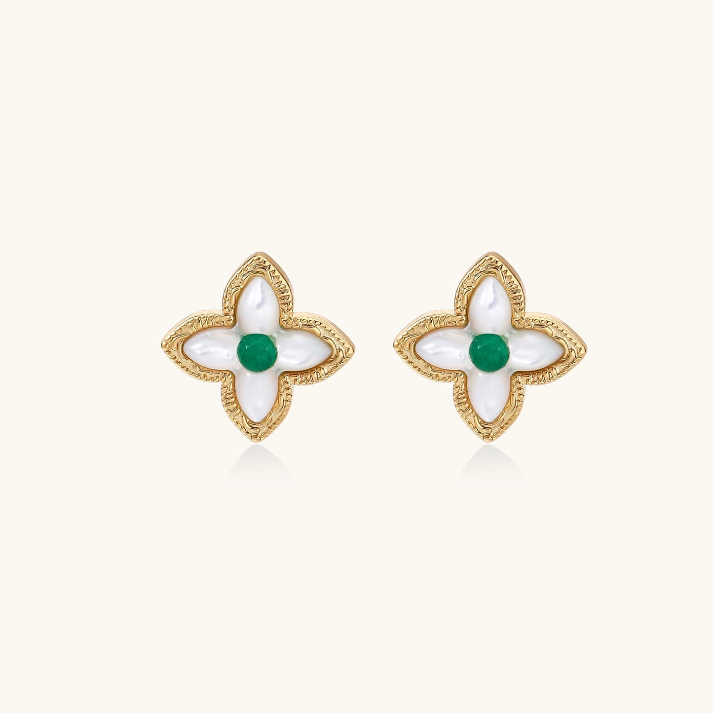 Dainty gold-plated quatrefoil stud earrings with white mother-of-pearl petals and green gemstone centers on beige background.