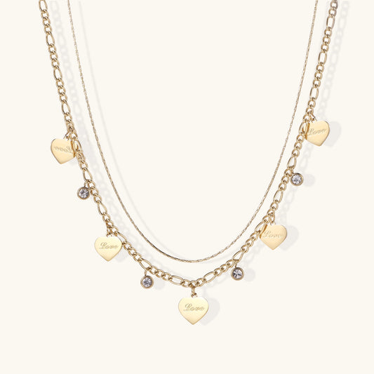 Two layered gold-plated necklaces with heart pendants engraved 'Love' and sparkling crystal charms on white background.