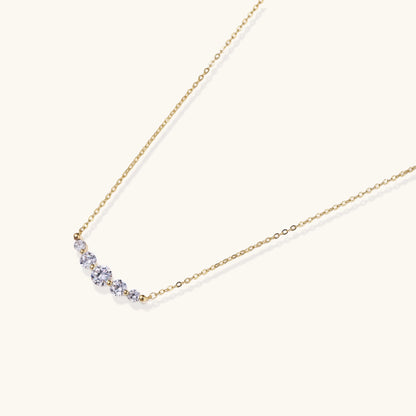 Delicate 14K gold plated necklace with curved arc of sparkling zirconia stones on cream background.