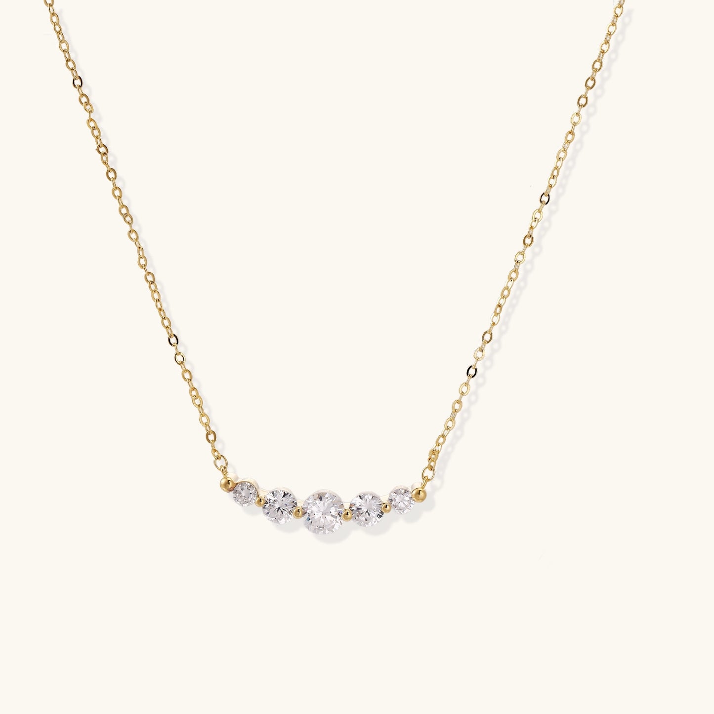 Delicate 14K gold plated necklace with curved arc of sparkling zirconia stones on cream background.