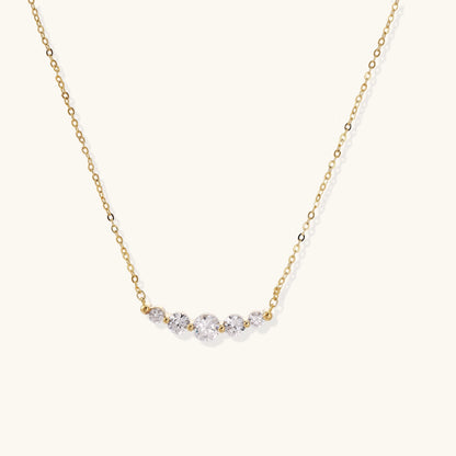 Delicate 14K gold plated necklace with curved arc of sparkling zirconia stones on cream background.