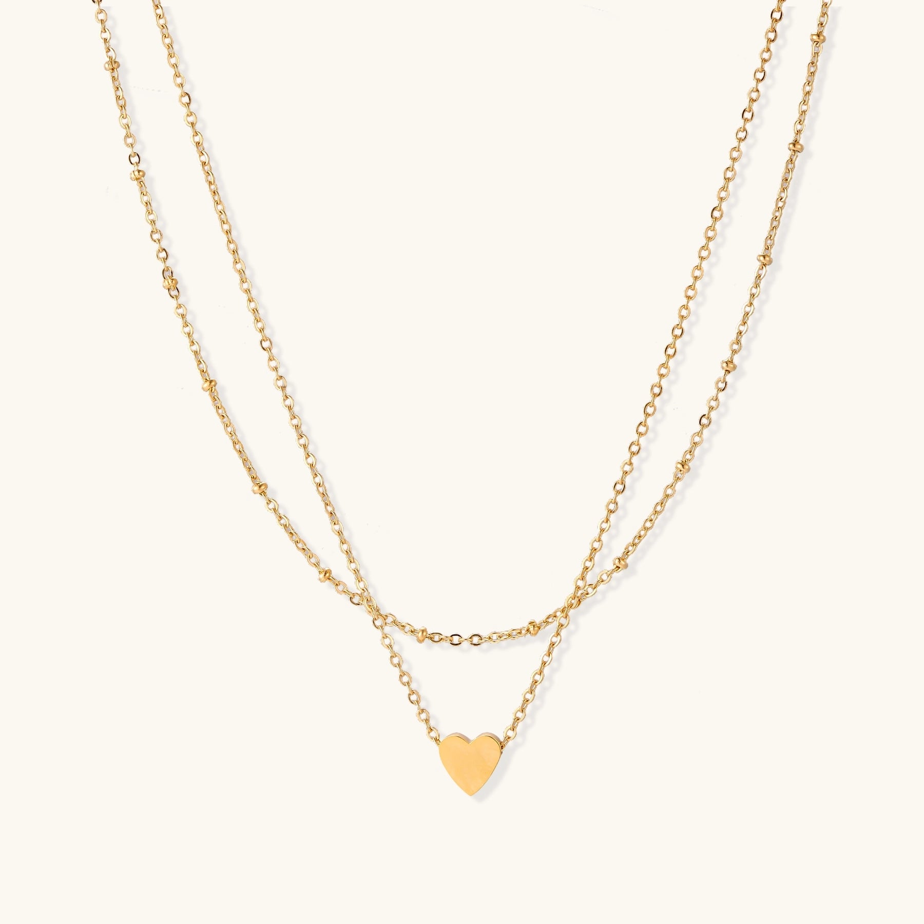 Two-layer gold-plated stainless steel necklace with small heart pendant displayed on beige background