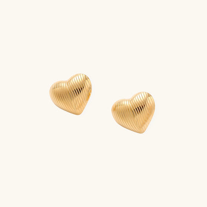 Close-up of ridged heart-shaped gold-plated stud earrings with textured surface pattern on cream background.
